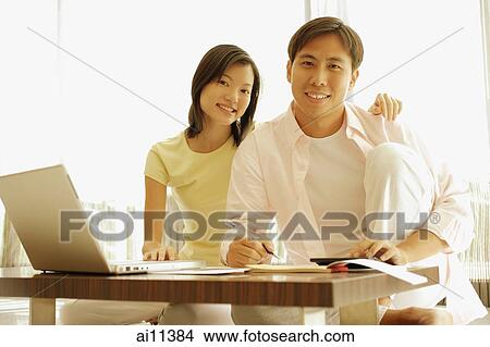 Couple sitting side by side at home, laptop open in front of them View Large Photo Image Picture - Couple sitting side by side at home, laptop open in front of them. Fotosearch