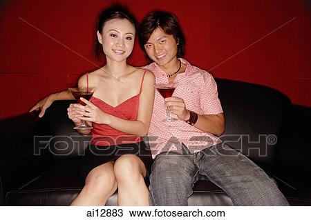 Stock Image - Couple sitting side by side, holding drinks, looking at camera. Fotosearch