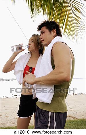 Stock Image - Couple standing at beach, side by side, woman drinking water. Fotosearch