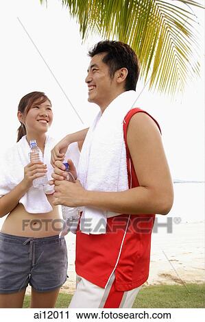 Stock Image - Couple standing at beach, side by side, holding water bottles. Fotosearch
