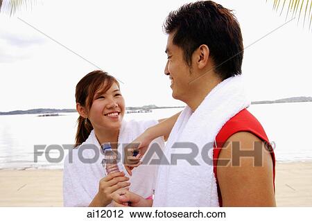 Stock Image - Couple standing at beach, side by side, holding water bottles, smiling. Fotosearch