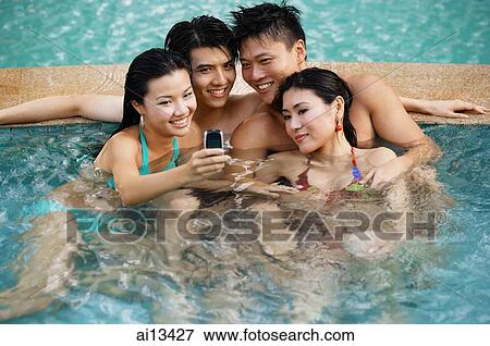 Stock Photo - Couples in swimming pool, looking at mobile phone, smiling. Fotosearch