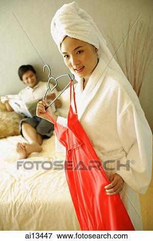 Stock Photo - Man lying on bed, woman in robe holding red dress in front of her. Fotosearch