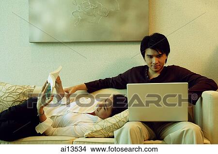 Man sitting on sofa, using laptop, woman lying next to him reading magazine View Large Photo Image Picture - Man sitting on sofa, using laptop, woman lying next to him reading magazine. Fotosearch