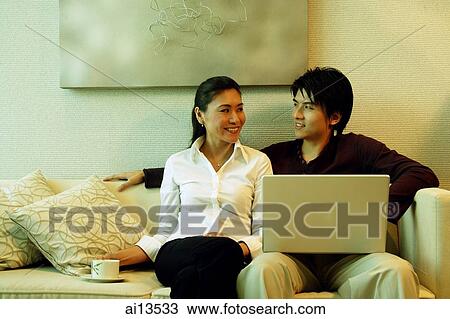 Stock Image - Man sitting on sofa, using laptop, woman sitting next to him. Fotosearch