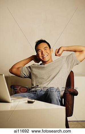 Stock Image - Man stretching while sitting on chair, laptop open in front of him. Fotosearch