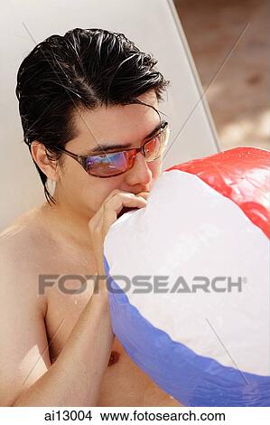 Picture - Man wearing sunglasses, blowing air into inflatable ball. Fotosearch
