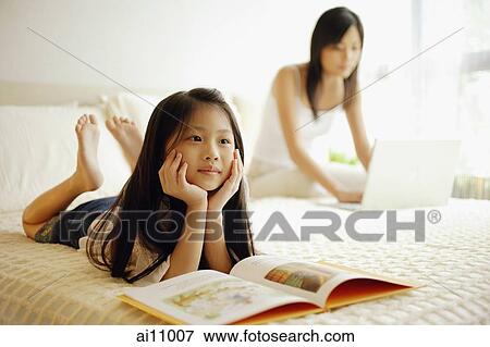 Stock Photo - Mother and daughter in bedroom, mother using laptop, daughter lying down with book open in front of her. Fotosearch