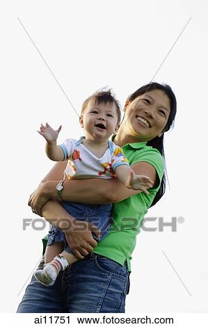 Stock Image - Mother carrying baby girl, smiling, low angle view. Fotosearch