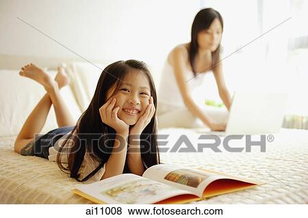 Mother using laptop, daughter lying down with book open in front of her View Large Photo Image Stock Photo - Mother using laptop, daughter lying down with book open in front of her. Fotosearch