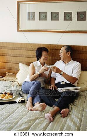 Stock Image - Older couple lying on bed, toasting with tea. Fotosearch