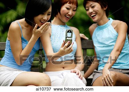 Stock Photo - Three young women sitting side by side, using camera phone. Fotosearch