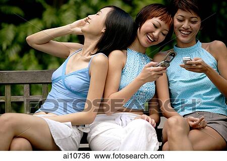 Stock Photograph - Three young women sitting side by side, using mobile phones. Fotosearch