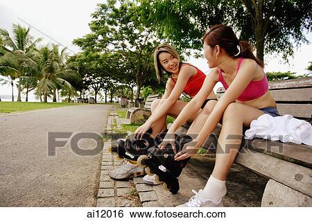 Two women sitting side by side on park bench, putting on roller blades View Large Photo Image Stock Photograph - Two women sitting side by side on park bench, putting on roller blades. Fotosearch