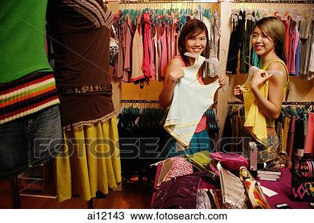 Two young women at clothes shop, holding up clothes, looking at camera View Large Photo Image Stock Image - Two young women at clothes shop, holding up clothes, looking at camera. Fotosearch