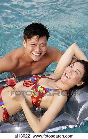 Stock Image - Woman on pool raft, looking at camera, man floating in water, next to her. Fotosearch