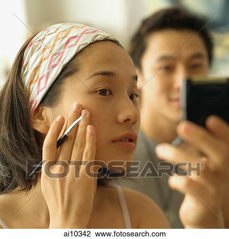 Stock Image - Woman touching face, looking at mirror, man in the background looking on.. Fotosearch