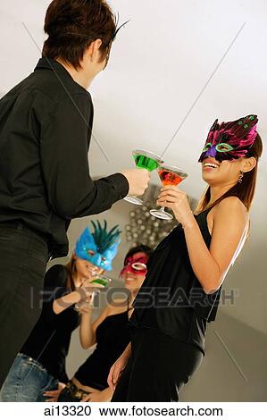 Stock Image - Young adults wearing masks and toasting with drinks. Fotosearch