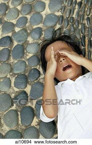 Young boy with hands over eyes, lying down on pebbled floor View Large Photo Image Picture - Young boy with hands over eyes, lying down on pebbled floor. Fotosearch