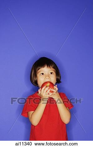 Stock Image - Young girl standing against blue background, biting an apple. Fotosearch