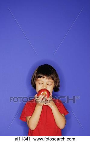 Young girl standing against blue background, holding an apple and biting it. View Large Photo Image Stock Image - Young girl standing against blue background, holding an apple and biting it.. Fotosearch
