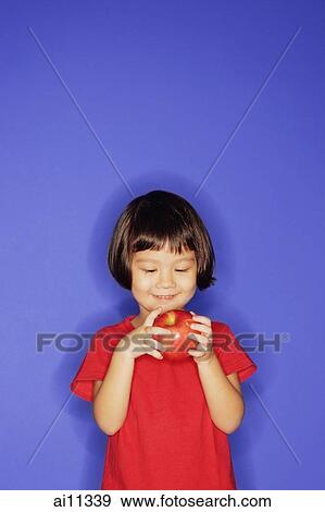 Stock Photo - Young girl standing against blue background, holding an apple. Fotosearch