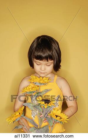 Stock Image - Young girl standing against yellow background, holding flowers. Fotosearch