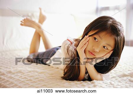 Stock Image - Young girl with hands on chin, lying on bed, looking away. Fotosearch