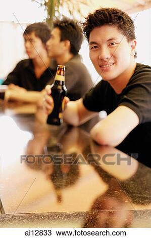 Young man at bar counter holding beer bottle, people in the background View Large Photo Image Stock Image - Young man at bar counter holding beer bottle, people in the background. Fotosearch