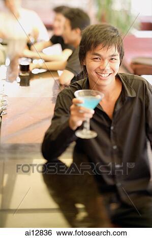 Young man at bar counter holding cocktail, people in the background View Large Photo Image Stock Photograph - Young man at bar counter holding cocktail, people in the background. Fotosearch
