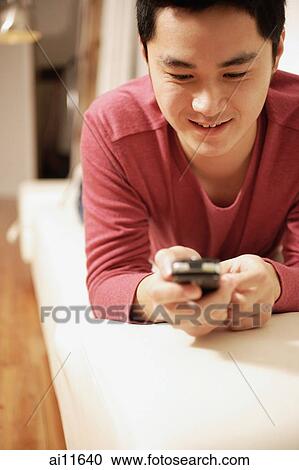 Stock Image - Young man holding mobile phone, lying on front. Fotosearch