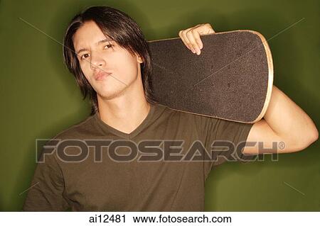 Stock Image -  Young man holding skateboard over shoulder, looking at camera. Fotosearch