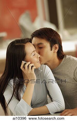 Stock Photograph - Young man kissing young woman, woman on the phone. Fotosearch