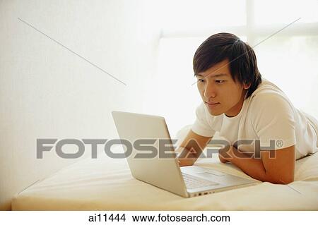Young man lying on bed, using laptop View Large Photo Image Picture - Young man lying on bed, using laptop. Fotosearch