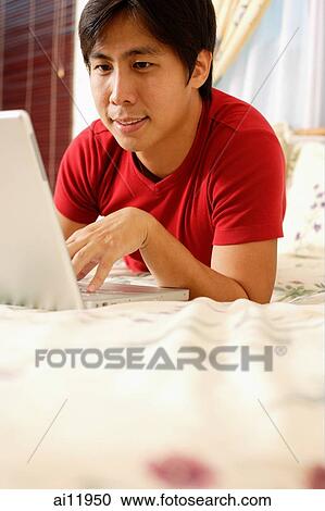 Young man lying on bed, using laptop View Large Photo Image Stock Image - Young man lying on bed, using laptop. Fotosearch