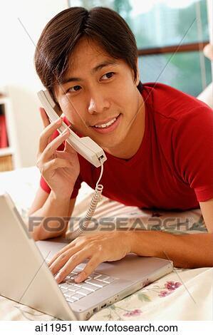 Young man lying on bed, using laptop and phone View Large Photo Image Stock Image - Young man lying on bed, using laptop and phone. Fotosearch