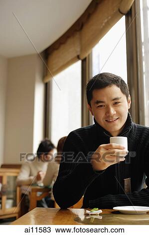 Stock Image - Young man sitting at table holding teacup, looking at camera. Fotosearch