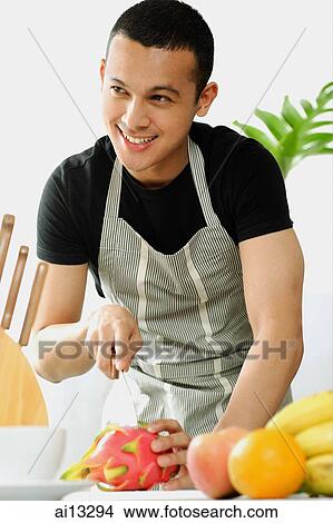 Picture - Young man wearing apron, cutting fruit, looking away, smiling. Fotosearch