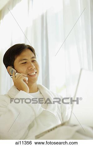 Stock Photograph - Young man wearing robe, using laptop and using mobile phone. Fotosearch