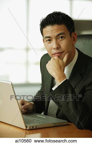 Young man with hand on chin, looking at camera, laptop open in front of him View Large Photo Image Stock Image - Young man with hand on chin, looking at camera, laptop open in front of him. Fotosearch