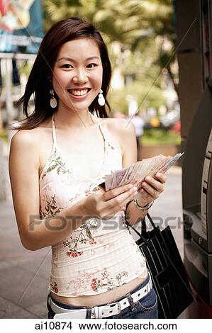 Picture - Young woman at an ATM machine, holding cash, looking at camera. Fotosearch
