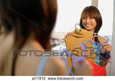 Young woman holding clothes, looking at mirror View Large Photo Image Stock Image - Young woman holding clothes, looking at mirror. Fotosearch
