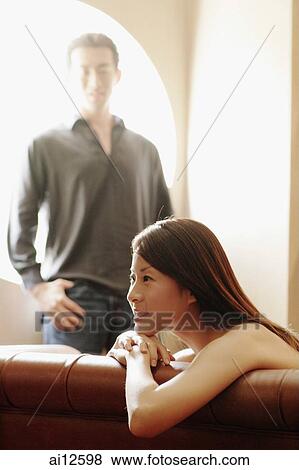 Young woman leaning over sofa, looking away, man in the background View Large Photo Image Stock Photo - Young woman leaning over sofa, looking away, man in the background. Fotosearch
