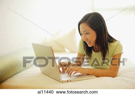 Young woman lying on bed, using laptop, smiling View Large Photo Image Stock Image - Young woman lying on bed, using laptop, smiling. Fotosearch