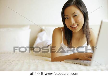 Young woman lying on bed, using laptop and smiling at camera View Large Photo Image Stock Photograph - Young woman lying on bed, using laptop and smiling at camera. Fotosearch
