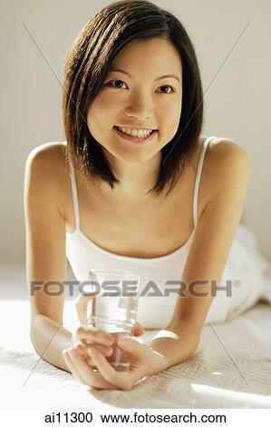 Stock Image - Young woman lying on front, holding glass of water. Fotosearch