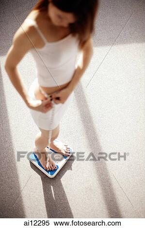 Young woman on weighing scale holding a tape measure around her waist View Large Photo Image Stock Photography - Young woman on weighing scale holding a tape measure around her waist. Fotosearch