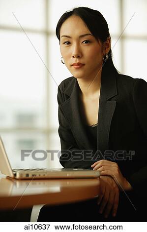 Stock Photo - Young woman sitting at desk, looking at camera, laptop open in front of her. Fotosearch