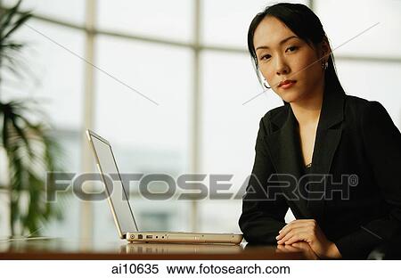Young woman sitting at desk, looking at camera, laptop open next to her View Large Photo Image Stock Photography - Young woman sitting at desk, looking at camera, laptop open next to her. Fotosearch