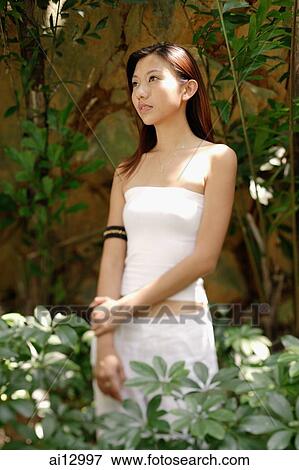 Stock Photo - Young woman standing and looking away, plants around her. Fotosearch